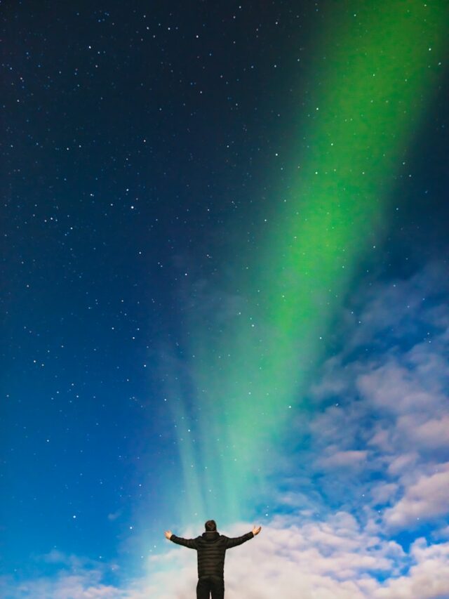 man standing on rock under green aurora borealis and white clouds